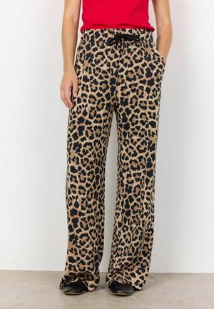 Person iført løse bukser med leopardprint med sort snøre og sorte sko, kombineret med en rød top, stående op ad en hvid væg.