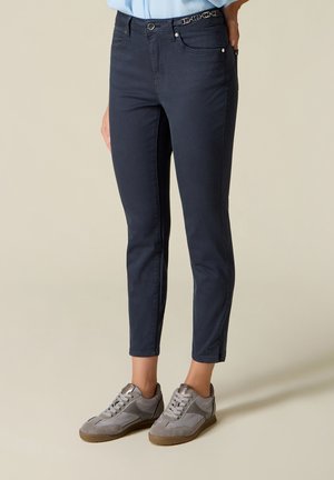 Kvinde iført marineblå slim-fit bukser med kædedetalje på taljen og grå snøresneakers, der står på en lys gulv.