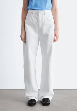 Osoba ubrana w białe szerokie spodnie z wysokim stanem, czarne trampki i jasnoniebieską koszulę włożoną do środka, stojąca na tle białej ściany.