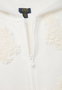 Sudadera blanca con cremallera, hecha de una tela suave, con aplicaciones florales en la parte delantera, un tirador de cremallera en forma de flor y una etiqueta de tela en el interior.