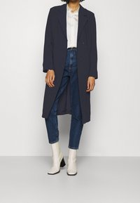 Cappotto lungo blu con una vestibilità ampia, abbinato a una blusa bianca e jeans blu a vita alta. Stivaletti bianchi con tacco completano il look.