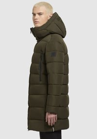 Puhasta jakna temno olivno zeleno, z kapuco, z zadrgo na žepih in prešito zasnovo z gladko teksturo. Črn logotip na rokavu.