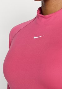 Hosszú ujjú rózsaszín sportfelső magas gallérral, sima anyagból készült. A mellkasi részen egy kis fehér Nike logó található.