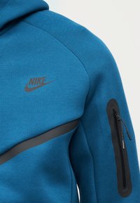 Zila Nike džemperis ar kapuci, kas izgatavots no teksturētas auduma, melniem akcentiem un rāvējslēdzēju kabatu uz piedurknes. Krūškurvī iespiests logotips.