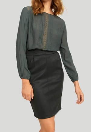 Vrouw draagt een donkergroene blouse met lange mouwen, kant en pintuckdetails, ingestopt in een hoog getailleerde zwarte kokerrok.
