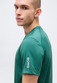 Νέος άνδρας με κοντά μαλλιά, ντυμένος με πράσινο αθλητικό μπλουζάκι που έχει την άσπρη επιγραφή "BORG" στο μανίκι, κοιτάζει πλάγια σε ένα απλό φωτεινό φόντο.