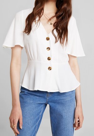 Vrouw draagt een witte blouse met korte mouwen met grote bruine knopen en een blauwe spijkerbroek, geaccentueerd met gelaagde gouden kettingen en een ring.