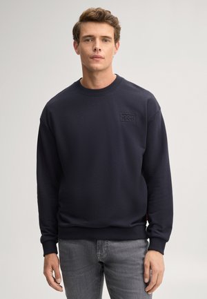 Uomo con una felpa blu navy a maniche lunghe con un piccolo logo "Joom Jeans" e jeans grigi, in piedi davanti a uno sfondo chiaro.