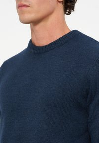 Homme portant un pull en maille bleu foncé à col rond, visible des épaules au menton sur fond uni.