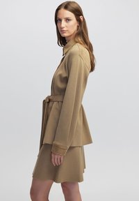 Abrigo envolvente color beige con cuello y mangas largas, hecho de material suave. Combinado con una falda acampanada a juego. Diseño simple y elegante.