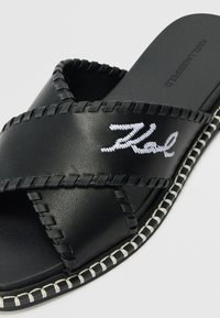 Black slide sandal with cross-stitched leather straps, white embroidered logo, and a textured footbed. Features a contrasting white stitched sole.