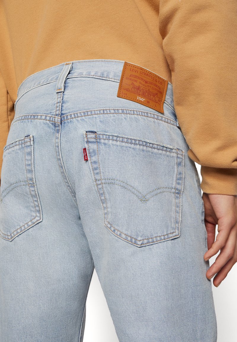 Hellblaue Jeans aus Denim haben ein braunes Lederpatch am hinteren Bund, kontrastierende Nähte und zwei hintere Taschen mit geschwungenem Design.
