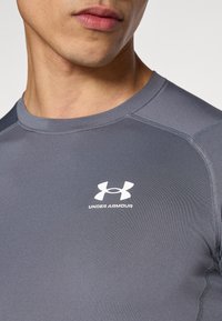 Γκρι αθλητικό μπλουζάκι από ελαστικό υλικό· διαθέτει στρογγυλή λαιμόκοψη, κοντά μανίκια και λευκό λογότυπο Under Armour στο στήθος.