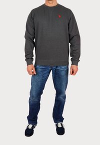 Grå sweatshirt med rund hals och röd logotyp, ihopparad med blå denimjeans och svarta sneakers med vita snören.