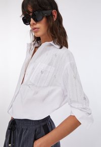 Chemise blanche boutonnée avec embellissements verticaux, une poche poitrine, manches retroussées, associée à un pantalon sombre à rayures et des lunettes de soleil.