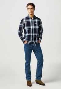 Camicia a quadri blu navy e grigia con bottoni, maniche lunghe, dettaglio sulla tasca anteriore, abbinata a jeans blu e stivali marroni. Tessuto dalla superficie liscia.