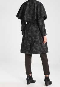 Černý a šedý vzorovaný dlouhý kabát s límcem ve stylu pláště, pásem v pase a kombinovaný s tmavými kalhotami a černými kotníkovými botami.