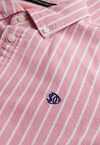 Camicia rosa con bottoni e righe verticali bianche, con logo ricamato blu navy sul petto.