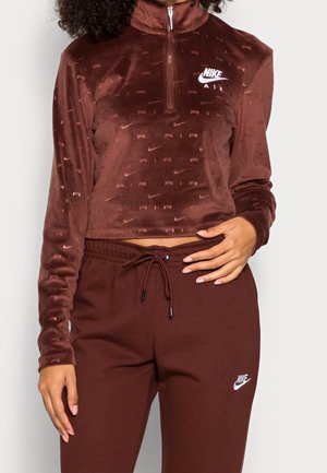 Kobieta w ciemnobrązowej krótkiej bluzie Nike Air na suwak oraz pasujących brązowych spodniach dresowych z białymi logo Nike.
