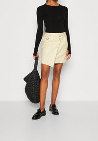 Haut noir à manches longues côtelé, jupe portefeuille crème texturée avec poches latérales, et sac noir tressé ; le mannequin porte des chaussures noires à lacets.