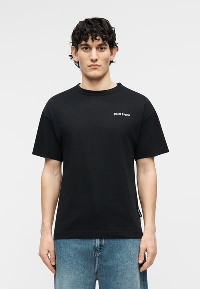 Νεαρός άνδρας με σγουρά σκούρα μαλλιά, που φοράει μαύρο T-shirt "Palm Angels" και μπλε τζιν, στέκεται μπροστά σε ένα απλό ανοιχτόχρωμο φόντο.