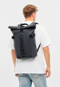 Mochila negra tipo roll-top con el logo OAK25, fabricada en material duradero, con correas ajustables y bolsillo lateral con cremallera, vista desde atrás.