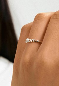 Anello d'argento con la parola "mom" in un design ondulato, posato su una mano con una texture della pelle liscia. Estetica semplice e moderna.