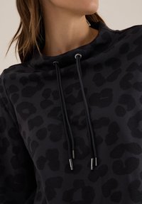 Hoodie noir avec un imprimé léopard subtil, doté d'un col montant, de cordons de serrage et d'embouts métalliques. Tissu doux avec une surface texturée.
