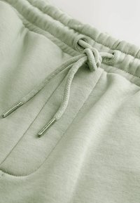 Taille de pantalon de survêtement vert clair avec cordon noué et embouts métalliques, montrant un tissu texturé et le détail de la couture de la poche.