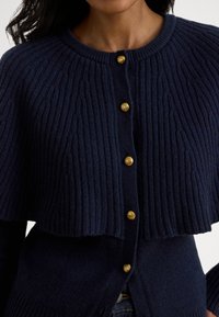 Tumesinist soonikuga kudumiga kampsunike, millel on ümar kaelus, silmatorkav tekstuur ja neli esiküljel asuvat kuldse tooni nööpi.