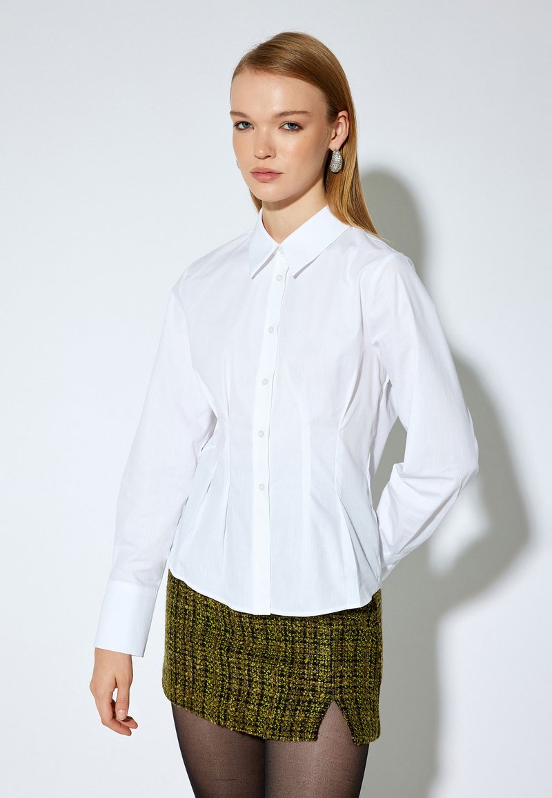 Camisa blanca de manga larga y botón con un diseño ajustado y cuello puntiagudo, combinada con una falda verde texturizada con una abertura lateral.