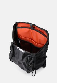 Bolso negro con forro naranja, que cuenta con múltiples compartimentos, un bolsillo de malla y correas ajustables. La tela presenta una superficie texturizada.