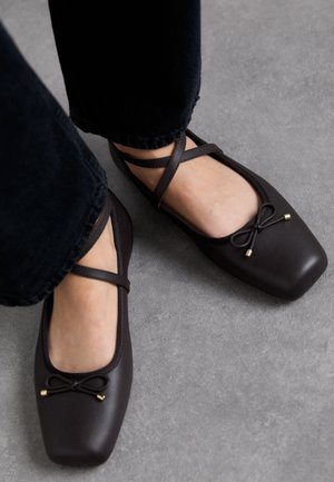 Feet wearing dark brown square-toe ballet flats with crisscross straps and small bow details, standing on gray floor.