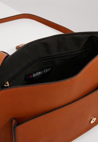 Hnědá kabelka z umělé kůže s černým vnitřkem, zipovou kapsou a značkovou visačkou. Má klopu a jednoduchý design popruhu.