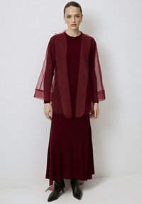 Abito a strati bordeaux con texture a coste, caratterizzato da una sovrapposizione trasparente e maniche svasate, abbinato a stivaletti neri a punta.