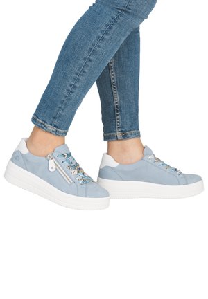 Persona che indossa sneaker celesti con suole bianche, lacci multicolore, cerniere laterali e jeans skinny blu.