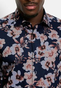 Marineblaues Blumenshirt mit großen hell- und dunkelrosa Blumen, subtilen Blättern, einem spitzen Kragen und zwei schwarzen Knöpfen an der Blende.