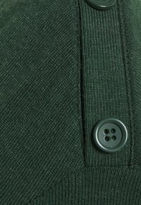 Tissu vert foncé texturé avec une couture visible et un bouton. Le bouton présente quatre trous et une surface lisse.