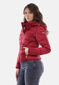 Giacca a vento rossa con zip frontale, cappuccio e motivo a rombi diagonali. Presenta tasche laterali e dettagli con logo sulla manica.