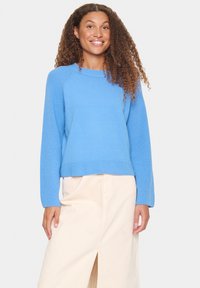Pull en tricot bleu clair avec des poignets et un bas côtelés, présentant une coupe décontractée. Le modèle l'associe à une jupe beige avec une fente à l'avant.