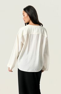 Long-sleeve blouse in soft, off-white fabric with a relaxed fit. Features a rounded hem and a subtle texture with visible seam detail.