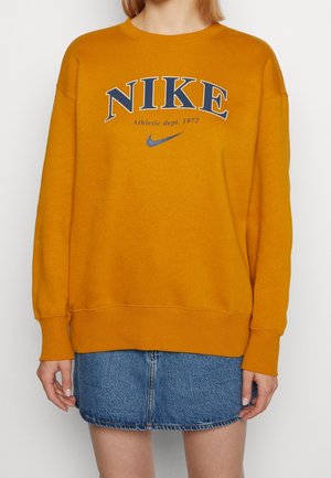 Personne portant un sweat-shirt Nike jaune moutarde avec logo et texte bleus, associé à une mini-jupe en denim bleu sur un fond clair uni.