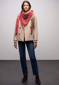 Beige dobbeltradet frakke med mørke knapper, båret over en sort top, kombineret med blå straight-leg jeans og brune ankelstøvler. Rødt og pink mønstret tørklæde.