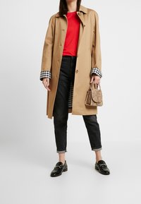 Cappotto trench beige con polsini a quadri bianco e nero, top rosso, jeans neri e mocassini neri. Porta una piccola borsa a tracolla con stampa serpente texturizzata.