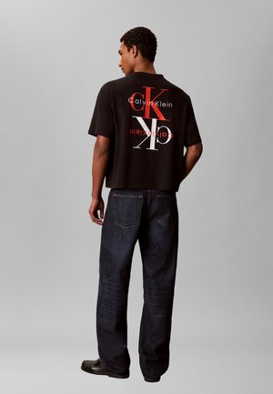 Mand iført sort Calvin Klein t-shirt med stort rødt og hvidt CK-logo og mørke jeans, stående med fronten væk fra kameraet mod ensartet grå baggrund.