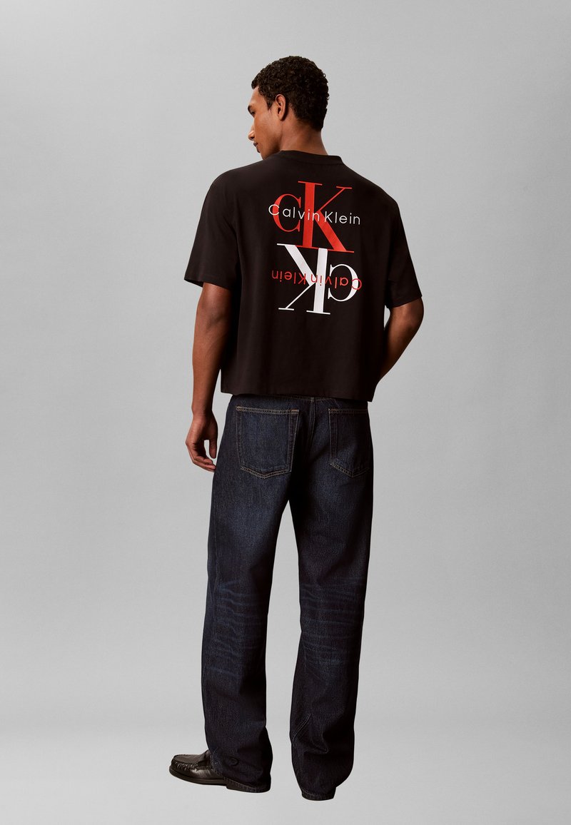 Άντρας φοράει μαύρο t-shirt Calvin Klein με μεγάλο κόκκινο και λευκό λογότυπο CK και σκούρο τζιν, στέκεται με την πλάτη προς τα εμπρός σε απλό γκρι φόντο.