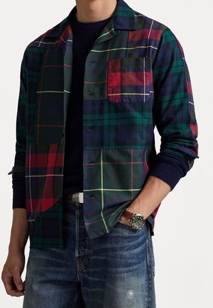Homme portant une chemise à carreaux boutonnée vert foncé, rouge et bleu marine sur un pull bleu marine, avec un jean bleu délavé et une montre-bracelet en cuir marron.
