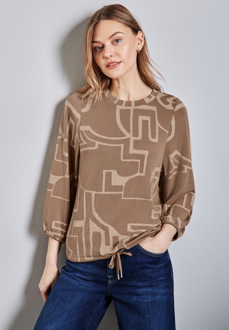 Brun långärmad topp med abstrakta beige mönster, rund halsringning och volangärmar. Matchad med mörkblå jeans.