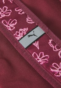 Tela marrón con un patrón floral rosa, que presenta una etiqueta gris con un logo negro de Puma. Textura suave con acentos en contraste.