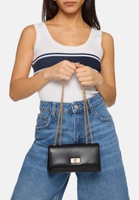 Borsa a tracolla in pelle nera con chiusura dorata e tracolla a catena, forma rettangolare, texture liscia, tenuta da una persona che indossa una maglietta bianca e jeans blu.